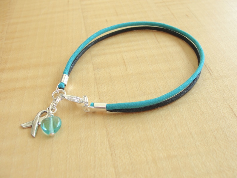 May include: A teal and black braided bracelet with a silver charm featuring a teal heart and a silver ribbon.