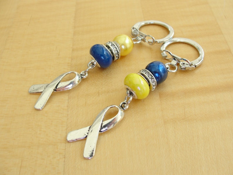 May include: Two silver keychains with blue and yellow beads and silver ribbon charms. The keychains are on a wooden surface.