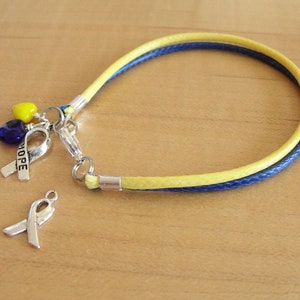 May include: A yellow and blue braided bracelet with a silver charm featuring a ribbon with the word "HOPE" and two small glass beads, one yellow and one blue.