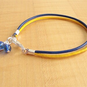 May include: A handmade bracelet featuring a navy blue and yellow leather band. The bracelet has a silver clasp and is adorned with two blue glass beads and a small silver charm. The bracelet is shown on a light brown wooden surface.