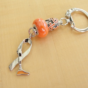 May include: Orange and silver charm keychain with a ribbon and a prosthetic leg. The charm is attached to a silver ring.