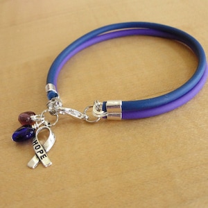 May include: A purple and blue rubber bracelet with a silver charm featuring a ribbon with the word "HOPE" engraved on it. The charm also has three small glass beads in shades of purple, blue, and red.