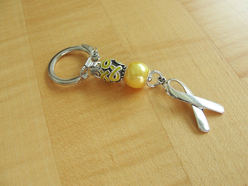 May include: A silver keychain with a yellow bead and a silver ribbon charm. The bead has a pattern of yellow and white swirls. The ribbon charm is in the shape of a ribbon.