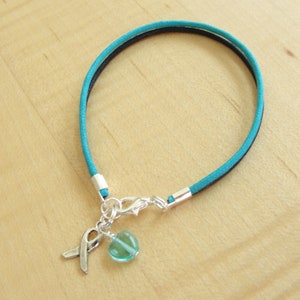 May include: Teal blue suede cord bracelet with a silver charm featuring a teal blue glass bead and a silver ribbon.