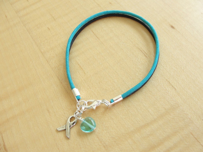 May include: Teal blue suede cord bracelet with a silver charm featuring a teal blue glass bead and a silver ribbon.