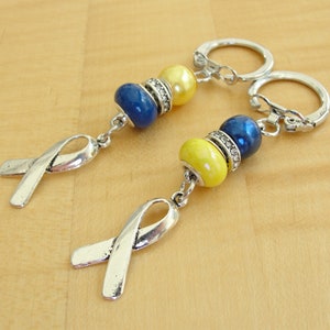 May include: Two silver keychains with blue and yellow beads and silver ribbon charms. The keychains are on a wooden surface.