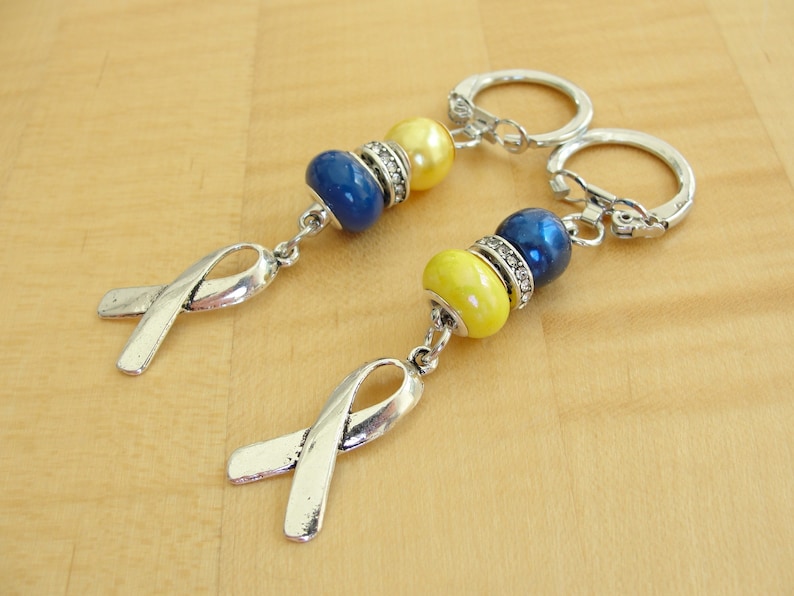 May include: Two silver keychains with blue and yellow beads and silver ribbon charms. The keychains are on a wooden surface.