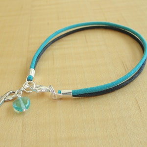 May include: A turquoise and black cord bracelet with a silver charm featuring a turquoise heart and a ribbon.