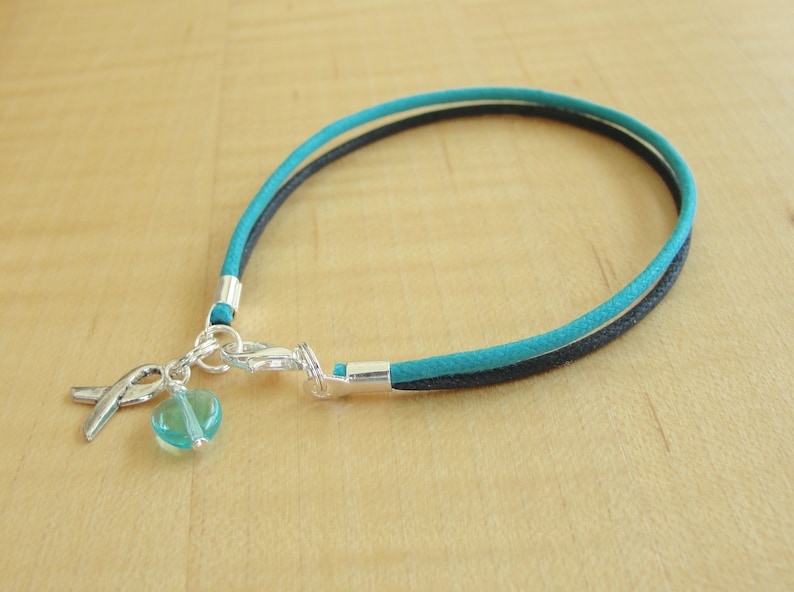 May include: A turquoise and black cord bracelet with a silver charm featuring a turquoise heart and a ribbon.