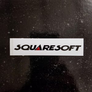 May include: A white rectangular sticker with the word "SQUARESOFT" in black, with a red triangle in the center of the word "SQUARE". The sticker is on a black background with white speckles.