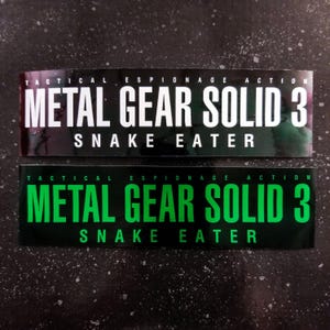 May include: Two rectangular stickers featuring the text "METAL GEAR SOLID 3 SNAKE EATER". The top sticker has white text on a black background. The bottom sticker has green text on a black background. The words "TACTICAL ESPIONAGE ACTION" are above the main text.