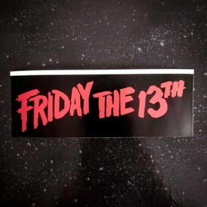 Può includere: Un adesivo rettangolare con la scritta "FRIDAY THE 13TH" in rosso. Il testo è su sfondo nero, con un bordo bianco in alto. L'adesivo è probabilmente per i fan del franchise di film horror.