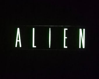 Puede incluir: Un letrero rectangular que presenta la palabra ALIEN en letras mayúsculas. El texto está iluminado en un color verde pálido brillante sobre un fondo negro sólido. La fuente es delgada, sin remates y espaciada uniformemente.