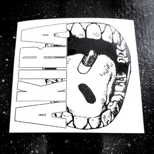 Op de afbeelding: Een zwart-witte sticker met het woord "AKIRA" in vette, omrande letters. Het ontwerp bevat een gedetailleerde illustratie van een open mond met tanden, een tong en twee pillen.