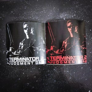 May include: Two open booklets featuring the Terminator 2: Judgment Day movie poster. One is black and white, the other red and black. The image shows a man with sunglasses holding a weapon. The movie title is at the bottom.