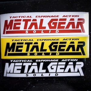 May include: Three rectangular stickers with the text "TACTICAL ESPIONAGE ACTION" and "METAL GEAR SOLID". The top sticker has a red background with white text, the middle one is yellow with black text, and the bottom one is black with white text.
