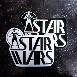 May include: Two stickers with the text "STAR WARS" in bold, white letters. One sticker is rectangular with a black background, and the other is a rounded rectangle with a white background. A circular graphic of a figure holding a weapon is above the text.