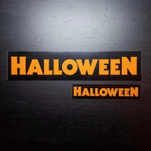 Può includere: Due adesivi a tema Halloween. L'adesivo più grande presenta la parola "HALLOWEEN" in lettere arancioni su sfondo nero. L'adesivo più piccolo dice anche "HALLOWEEN" in lettere arancioni su sfondo nero.