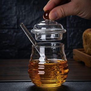 May include: A clear glass honey pot with a lid and a honey dipper. The pot is filled with golden honey. The lid is being lifted by a hand. The pot has a ribbed design around the base.