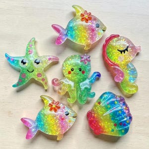 Könnte beinhalten: Eine Sammlung farbenfroher, glitzernder Meereskreaturen-Ornamente, darunter Fische, ein Seestern, ein Oktopus, ein Seepferdchen und eine Muschel. Jedes Ornament hat einen Regenbogeneffekt und ist mit kleinen Blumen verziert.