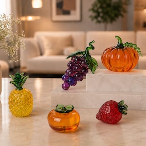 May include: A selection of glass fruit ornaments, featuring a pineapple, grapes, pumpkin, orange, and strawberry. The pineapple is yellow, the grapes are purple, the pumpkin and orange are orange, and the strawberry is red. Each piece has detailed craftsmanship.