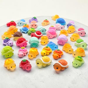 May include: A selection of small, brightly coloured turtle figurines. The turtles are in shades of yellow, orange, pink, blue, and green. Some have fruit-themed shell designs, like strawberries and grapes. The figurines are small and seem to be made of a translucent material.