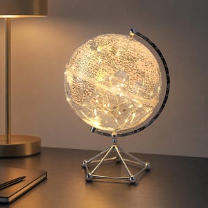 May include: A clear globe featuring a world map, lit with warm white fairy lights. The globe sits on a silver-coloured metal stand. A gold-coloured lamp is visible in the background.