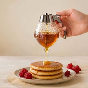 Puede incluir: Un dispensador de miel de vidrio con un patrón de panal vierte miel sobre una pila de panqueques. El dispensador tiene una tapa y un asa plateadas. Las frambuesas frescas están en un plato junto a los panqueques. La miel es de color ámbar dorado.