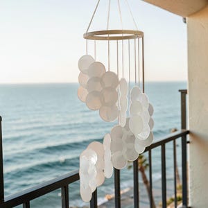 Può includere: Una carillon eolico di conchiglie capiz bianche con una parte superiore circolare e più fili di dischi di conchiglia. Il carillon è appeso a un filo sottile, con l'oceano visibile sullo sfondo. Le conchiglie sono traslucide e disposte in grappoli a cascata.