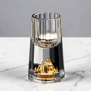 May include: A clear glass tumbler with a gold mountain design at the base. The upper portion of the glass has vertical ridges. The base of the glass is black and clear, sitting on a white surface.