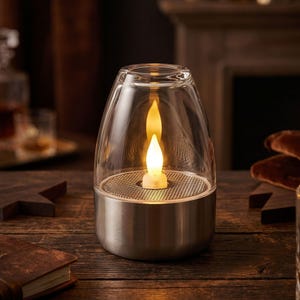 May include: A glass lantern with a silver base, housing a flickering LED candle. The lantern is set on a wooden table. The background is blurred, showing a fireplace and other decorative items.
