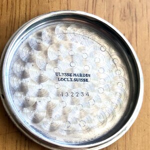 May include: Close-up of the inside of a silver-colored watch case. The text "ULYSSE NARDIN LOCLE SUISSE" is engraved at the top, with the number "432234" below it. The interior has a textured, circular pattern.