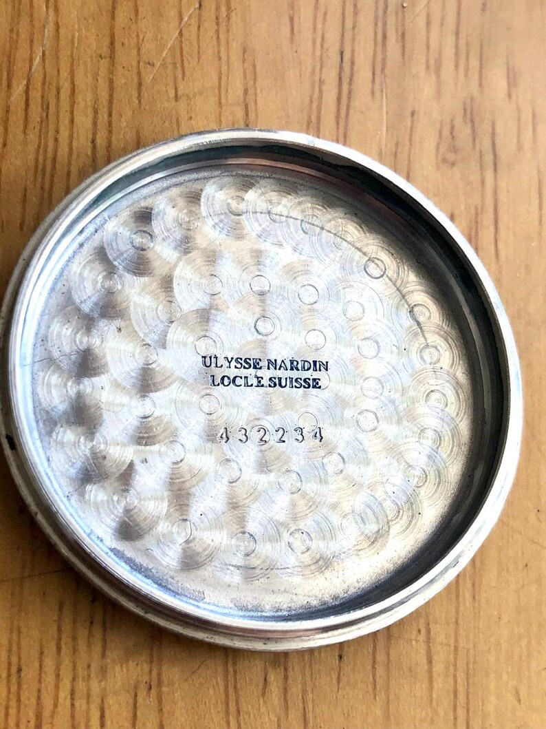 May include: Close-up of the inside of a silver-colored watch case. The text "ULYSSE NARDIN LOCLE SUISSE" is engraved at the top, with the number "432234" below it. The interior has a textured, circular pattern.