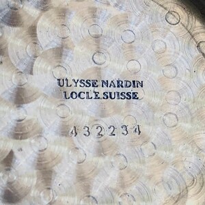 May include: Close-up of a watch back with a textured, silver-colored surface. The text "ULYSSE NARDIN LOCLE SUISSE" is engraved in black, along with the number "432234". The watch back has a circular design.