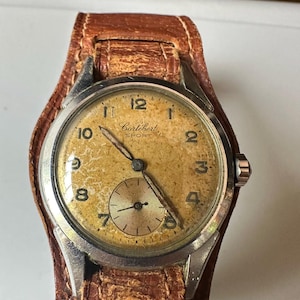 May include: Vintage Cortebert Sport wristwatch with a light brown face, silver case, and a small seconds dial. The watch has black numerals and hands, and is set on a wide, distressed brown leather strap.