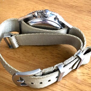 May include: A silver-toned wristwatch with a light green canvas band. The watch face has a black bezel and a silver case. The band is secured with a silver buckle. The watch is shown on a wooden surface.