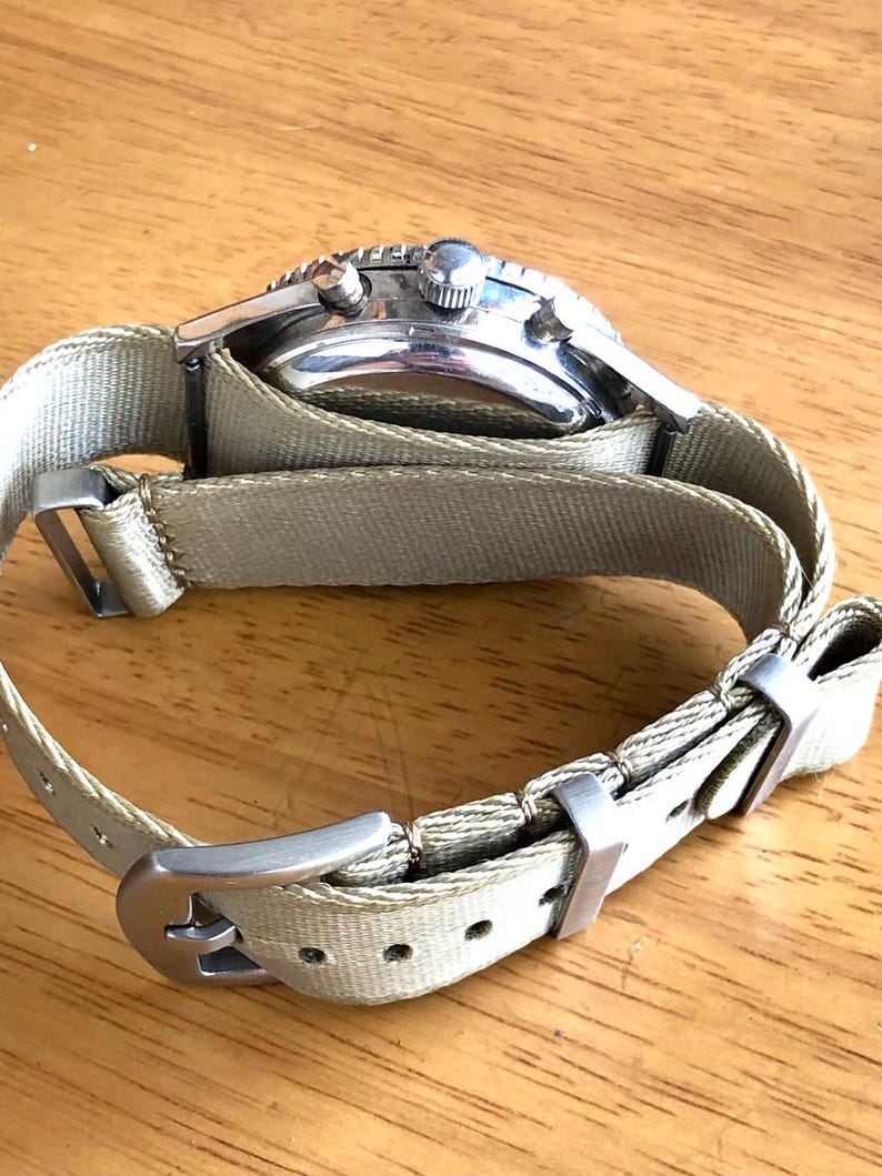 May include: A silver-toned wristwatch with a light green canvas band. The watch face has a black bezel and a silver case. The band is secured with a silver buckle. The watch is shown on a wooden surface.