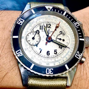 May include: Vintage chronograph watch with a cream-colored face, black bezel, and silver case. The watch features multiple sub-dials, luminous hands, and a fabric strap. The bezel has white numerals and markers. The watch is a classic timepiece.