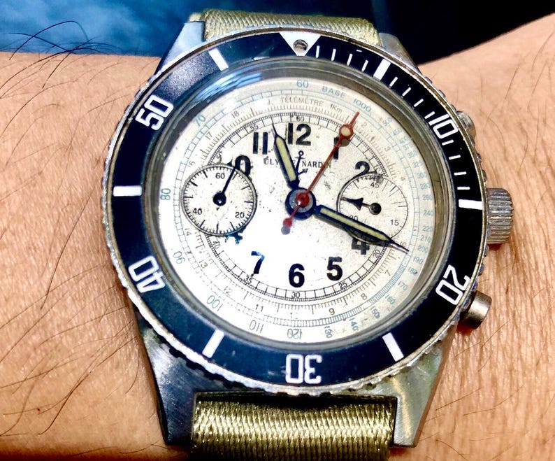May include: Vintage chronograph watch with a cream-colored face, black bezel, and silver case. The watch features multiple sub-dials, luminous hands, and a fabric strap. The bezel has white numerals and markers. The watch is a classic timepiece.