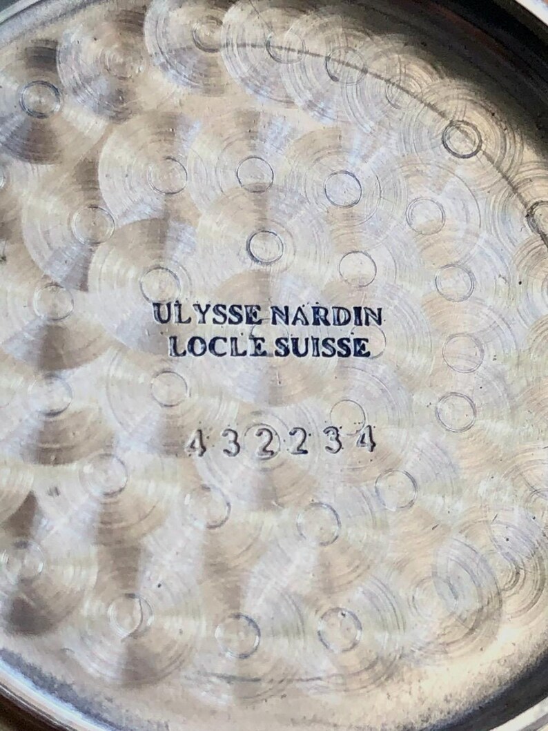 May include: Close-up of the back of a watch with a textured, silver-colored surface. The text "ULYSSE NARDIN LOCLE SUISSE" is engraved at the top, with the number "432234" below. The watch's design features concentric circles.