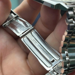 以下が含まれることがあります： 金属バンドが付いたシルバーのステンレススチール製腕時計のクローズアップ。クラスプが開いており、「STL ALL STAINLESS STEEL」の文字が見えます。背景には時計の文字盤が見えます。