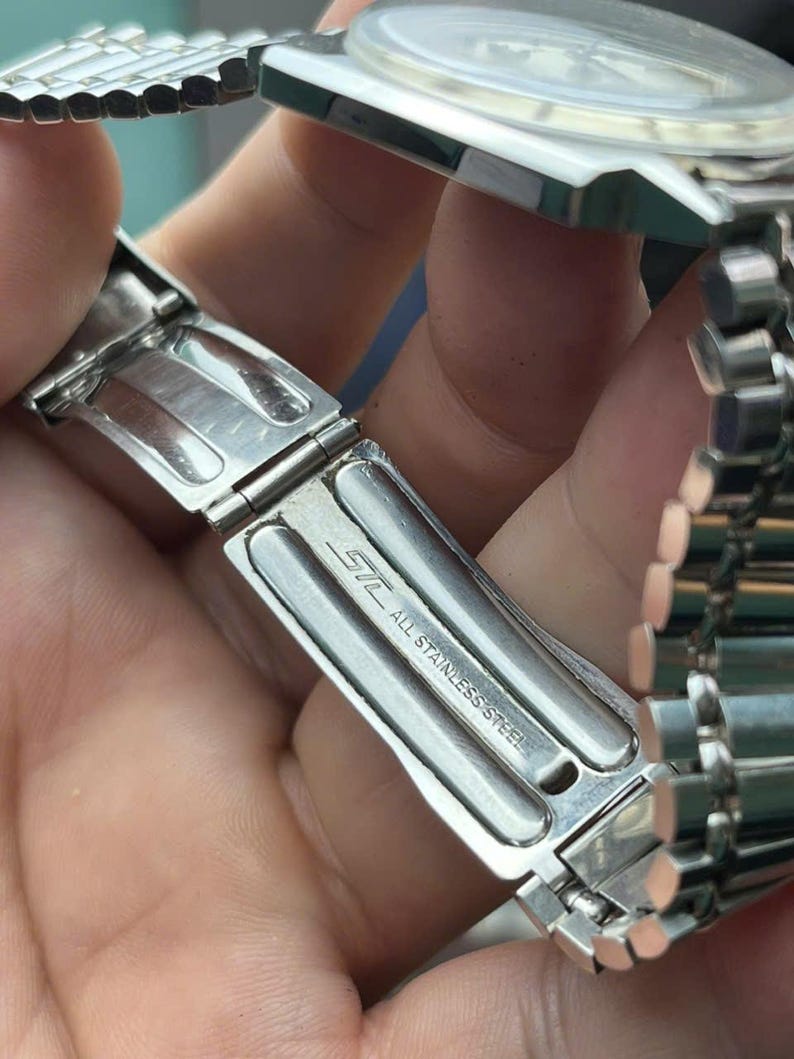 以下が含まれることがあります： 金属バンドが付いたシルバーのステンレススチール製腕時計のクローズアップ。クラスプが開いており、「STL ALL STAINLESS STEEL」の文字が見えます。背景には時計の文字盤が見えます。