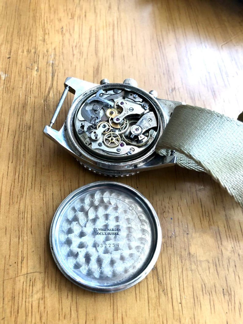 May include: A vintage Ulysse Nardin watch with the back removed, revealing the intricate internal mechanisms. The watch has a silver-colored case and a light green fabric strap. The watch's back is also visible, with the text "Ulysse Nardin Locle Suisse" and the number "432234".