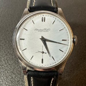 May include: A vintage wristwatch with a silver face and black hands. The watch has a silver case and a black leather strap with white stitching. The watch face displays the text "International Watch Co SCHAFFHAUSEN".