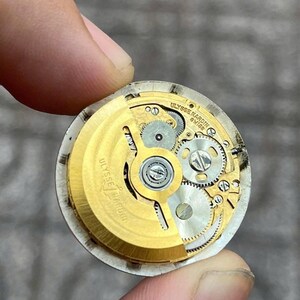 以下が含まれることがあります： 金色の歯車と「ULYSSE NARDIN SWISS」の文字が入った時計のムーブメントのクローズアップ。指で持たれており、時計の複雑な内部コンポーネントが写っています。