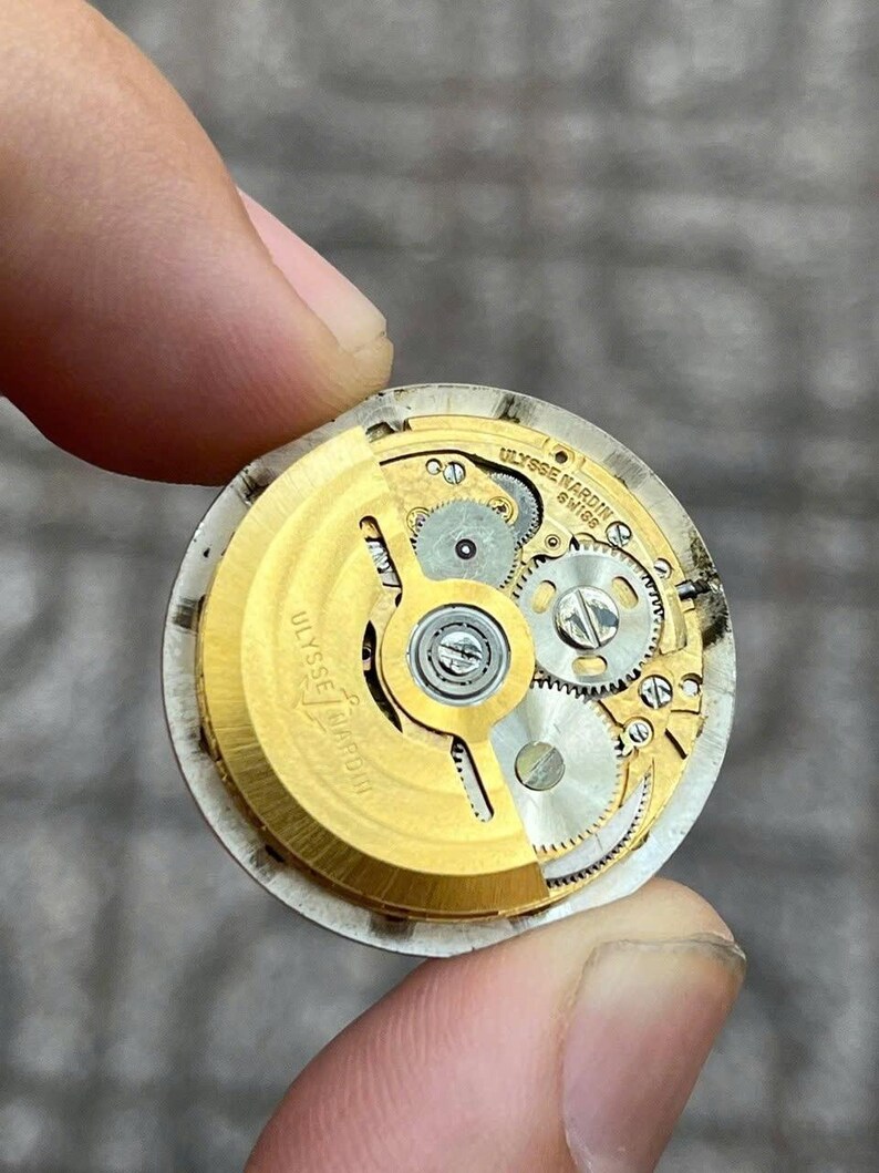 以下が含まれることがあります： 金色の歯車と「ULYSSE NARDIN SWISS」の文字が入った時計のムーブメントのクローズアップ。指で持たれており、時計の複雑な内部コンポーネントが写っています。