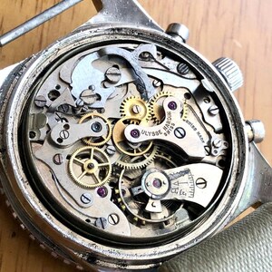 May include: Close-up of a vintage Ulysse Nardin Swiss watch mechanism. The watch's intricate internal gears, springs, and jewels are visible. The watch has a silver-colored case and a light-colored canvas strap.
