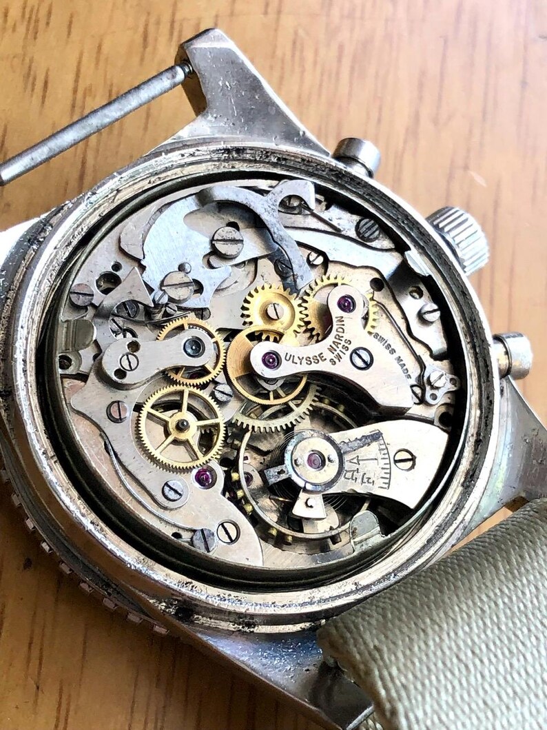 May include: Close-up of a vintage Ulysse Nardin Swiss watch mechanism. The watch's intricate internal gears, springs, and jewels are visible. The watch has a silver-colored case and a light-colored canvas strap.
