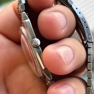 以下が含まれることがあります： 金属バンドが付いたシルバーカラーの腕時計。時計の文字盤が見え、リューズには装飾的なエンブレムがあります。手で持たれており、バンドのリンクと時計全体のデザインが示されています。