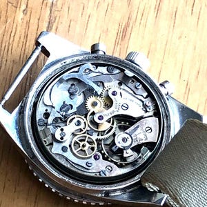 May include: Close-up of a vintage wristwatch's intricate internal mechanism. The watch features numerous small gears, jewels, and other components. The watch has a silver case and a green fabric strap. The watch is on a wooden surface.